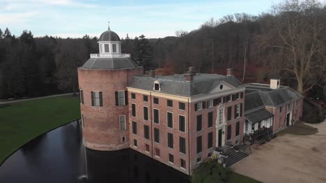 Slow-aerial-panning-around-exterior-facade-with-rook-tower-and-fountain-in-still-water-around-Rosendael-castle-and-garden-with-barren-winter-trees-in-the-background