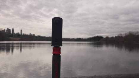 360-camera-on-monopod-on-the-edge-of-a-lake-capturing-landscape-photography-videography-of-water-clouds-trees-sky