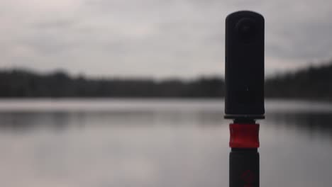 360-camera-on-monopod-on-the-edge-of-a-lake-capturing-landscape-photography-videography-of-water-clouds-trees-sky