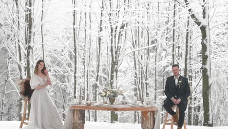 Cool-newlyweds-posing-by-table-in-snowy-winter-forest,-wedding-editorial