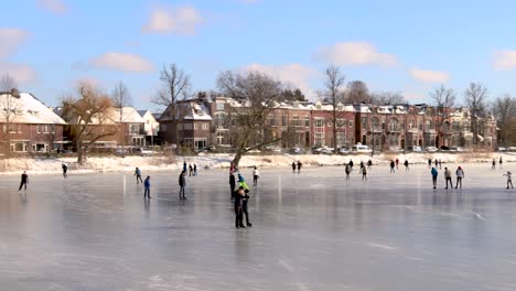 Ice-skating-people-on-picturesque-frozen-canal-in-city-environment-with-winter-barren-trees-and-white-snow-in-cold-cozy-landscape-including-former-water-tower-in-the-background