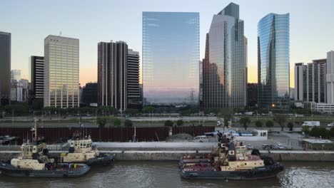Aerial-dolly-left-of-boats-in-Puerto-Madero-docks-near-Paseo-del-Bajo-highway-and-skyscrapers-at-sunset,-Buenos-Aires