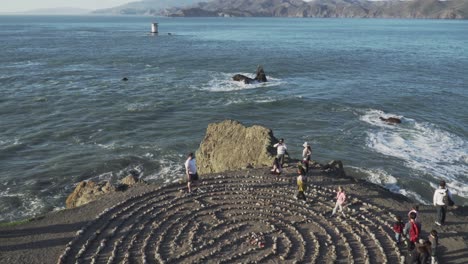 Tiro-Inclinado-De-Personas-Caminando-Por-El-Laberinto-De-Arte-Del-Laberinto-Del-Extremo-De-La-Tierra-Cerca-De-Las-Olas-Rompientes-De-La-Costa-Del-Pacífico