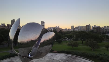 Dolly-in-flying-above-Floralis-Generica-steel-sculpture-with-Recoleta-buildings-in-background-at-golden-hour