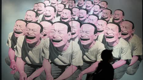 Visitors-wearing-face-masks-walk-past-Chinese-artist-Yue-Minjun-art-piece-named-"Self-portrait",-depicting-himself-while-grinning-with-his-mouth-gaping,-at-collectibles-Sotheby's-show-in-Hong-Kong