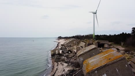 Aerial-view-of-abandoned-seaside-fortification-building-at-Karosta-Northern-Forts-on-the-beach-of-Baltic-sea,-Liepaja-in-overcast-spring-day,-wind-turbine-in-background,-wide-drone-shot-moving-forward