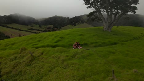 Man-With-Girlfriend-Sitting-On-Grass-Controlling-And-Flying-Drone-At-Purerua-Peninsula-On-Misty-Day-In-New-Zealand