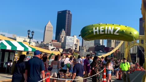 Menschen,-Die-Auf-Der-Andy-Warhol-Brücke-Laufen,-Mit-Einem-Riesigen-Heinz-Pickle-Ballon-Im-Hintergrund