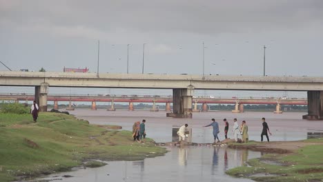 People-Help-Man-Cross-A-River-In-Pakistan,-Cars-Driving-On-Bridge-Overpass