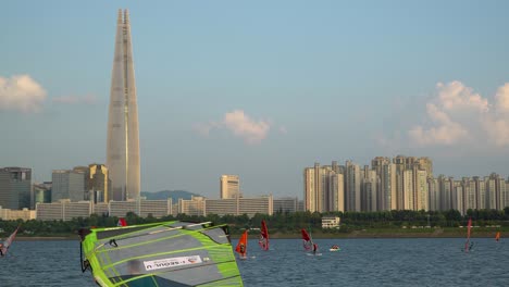 Windsurfistas-Surfeando-En-El-Río-Han,-Lotter-World-Tower-En-El-Fondo-Al-Atardecer,-Jamsil,-Seúl,-Corea-Del-Sur