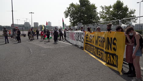 A-unit-of-police-officers-walk-past-protestors-holding-large-banner-that-says,-“UK-Stop-Arming-Israel”and-“Elbit-weapons,-tested-on-Palestinians”-at-a-protest-against-the-DSEi-arms-fair