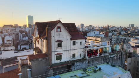 Panorámica-Aérea-Cinematográfica-Alrededor-Del-Pequeño-Chalet-Aislado-Muebleria-Diaz-Encaramado-En-La-Terraza-De-La-Azotea,-Con-Función-De-Puertas-Abiertas-Celebrada-En-El-Lugar-Con-El-Paisaje-Urbano-Del-Centro-Y-Una-Hermosa-Vista-Del-Atardecer