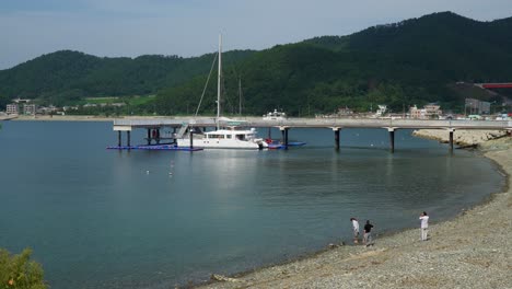 Catamaran-Boat-Docked-By-The-Jetty-With-Family-Relaxing-On-Pebble-Beach