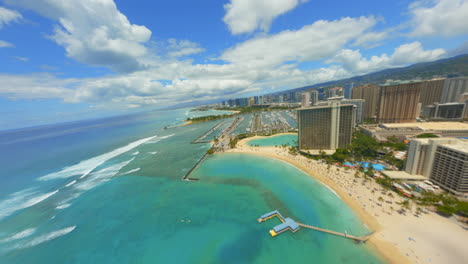 Volando-Sobre-Waikiki,-Fpv-Drone-Disparó-Sobre-La-Playa-De-Waikiki-Y-Hoteles,-Agua-Azul-Y-Surfistas-Con-Honolulu-En-La-Distancia