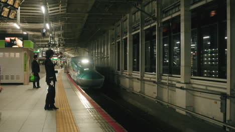 Night-shot-of-Japanese-bullet-train-approaching-train-platform-at-Sendai-Station,-Japan,-with-passengers-and-train-conductors-waiting-on-the-platform