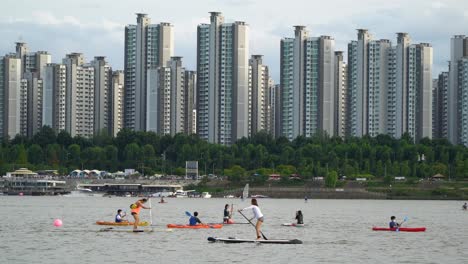 Gente-Coreana-Haciendo-Kayak,-Piragüismo,-Windsurf-Y-Remando-En-Tablas-De-Remo-De-Pie-Con-Edificios-Altos-De-Apartamentos-Jamsil-En-El-Fondo-Del-Río-Han,-En-El-Centro-De-Seúl,-Corea-Del-Sur