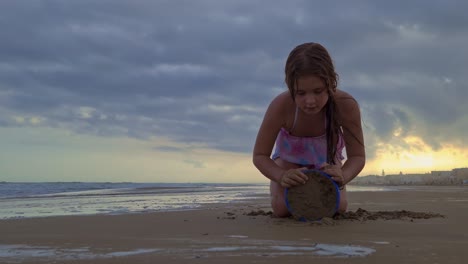 Low-angle-of-beautiful-caucasian-little-girl-playing-with-bucket-and-sand-on-beach-at-sunset