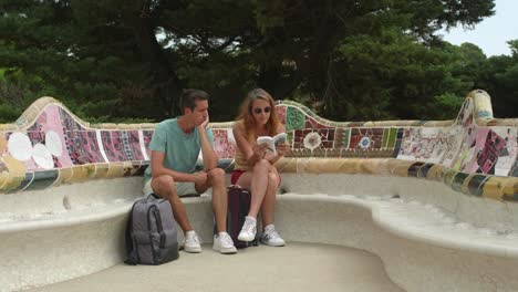 Ein-Junges-Paar,-Das-Auf-Den-Berühmten-Mosaik-Keramikbänken-Des-Park-Güell-Sitzt-Und-Seine-Reiseroute-Bespricht