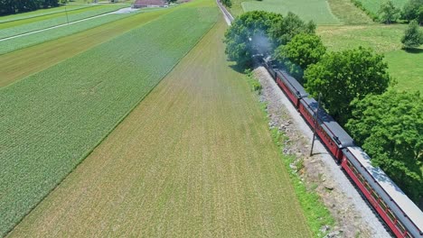An-Aerial-Overhead-View-of-a-Steam-Engine-Puffing-Smoke-and-Steam-with-Passenger-Coaches-Traveling-on-a-Single-Track-Thru-Planted-Fields-and-Farmland-Countryside-on-a-Beautiful-Cloudless-Spring-Day