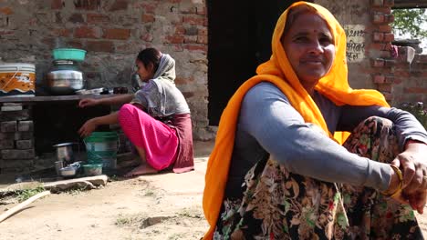 Mujer-India-Mirando-A-La-Cámara-Y-Su-Hija-Cocinando-Pan-Chapati-En-El-Fondo,-Rajasthan