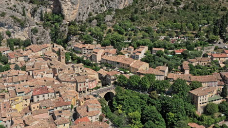 Moustiers-Sainte-Marie-France-Aerial-v7-establishing-shot,-birds-eye-view-drone-flyover-foothill-old-medieval-village,-tilt-up-reveals-spectacular-limestone-mountain-walls---July-2021