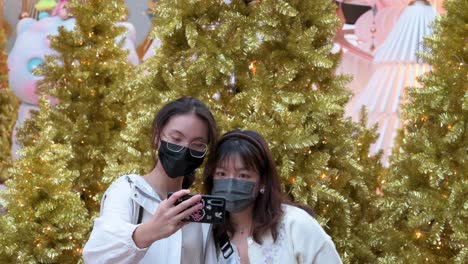 Freunde-Mit-Gesichtsmasken-Machen-Ein-Selfie-Mit-Goldenen-Weihnachtsbäumen-Im-Hintergrund,-Während-Sie-Ihren-Abend-Bei-Einer-Installationsveranstaltung-Zum-Thema-Weihnachten-In-Hongkong-Genießen