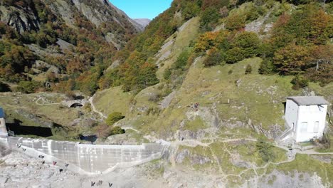 Lac-d'Oô-artificial-lake-in-the-French-Pyrenees-with-hikers-on-the-dam-wall-and-man-walking-dog,-Aerial-dolly-out-shot