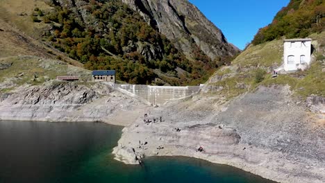 Lac-d'Oô-lake-in-the-French-Pyrenees-with-visitors-exploring-the-dam-wall-on-cliffside,-Aerial-orbit-around-shot