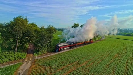 Vista-Aérea-De-Un-Motor-De-Vapor-Antiguo-Y-Autocares-De-Pasajeros-Que-Viajan-A-Lo-Largo-Del-Campo-Soplando-Humo-Y-Drones-Viajando-Paralelo-Y-Cerca-De-él,-En-Un-Día-Soleado-De-Verano