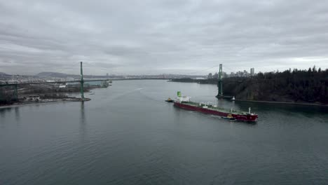 Red-barge-passing-under-Lions-Gate-bridge-with-Vancouver-city-in-background,-Canada