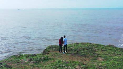 Toma-Aérea-Giratoria-De-Una-Pareja-Heterosexual-De-Pie-En-Un-Acantilado-Cubierto-De-Hierba-En-Los-Brazos-Del-Otro-Hablando-Y-Mirando-Al-Mar,-Día-Soleado,-Olas-Que-Reflejan-El-Sol,-El-Horizonte,-El-Mar-Azul,-El-Amor