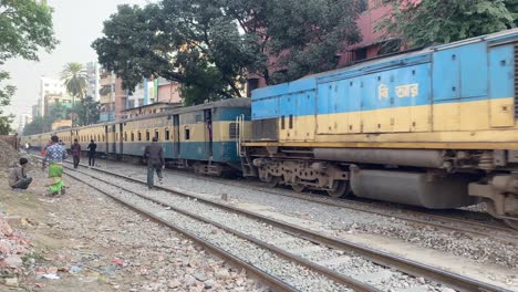 Bengalischer-Mann-Mit-Korb-Auf-Dem-Kopf,-Der-An-Bahngleisen-Vorbeigeht,-Mit-Einem-Zug-Im-Hintergrund-In-Dhaka