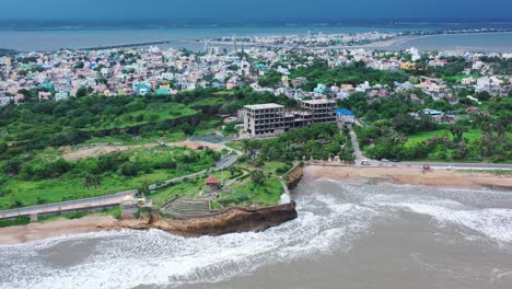 aerial-zoom-out-view-showing-various-buildings-and-greenery-right-on-the-coastline,-white-waves-gently-crashing-on-the-shore,-boats-by-the-skyline,-clear-blue-sky,-visible-horizon,-empty-roads,-city