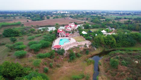 Aerial-surrounding-drone-view-of-private-double-pool-villa-in-Vadodara,-India,-with-a-blue-swimming-pool-and-surrounded-by-greenery-and-a-river