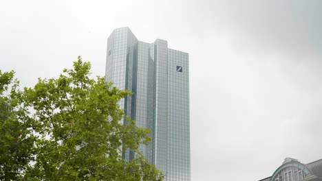 Low-shot-of-Deutsche-Bank-Tower-in-am-Main-Frankfurt-Germany-with-trees-in-the-foreground-and-a-bright-cloudy-sky