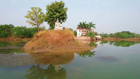Travel-across-the-lake-with-aerial-drone-in-a-beautiful-meditation-house-in-Vadodara,-India,-seeing-the-water,-the-Shiva-statue-and-the-luxurious-white-vacation-house-with-a-red-brick-roof