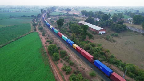 Freight-train-with-blue,-yellow,-red-and-light-blue-wagons-moving-through-the-green-harvest-fields-in-India-near-a-highway-on-a-cloudy-day