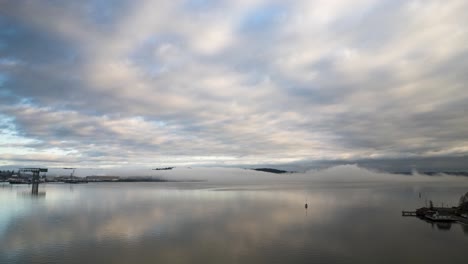 Fog-billows-across-still-water-as-marine-layer-clouds-stream-across-the-sky,-aerial-hyperlapse