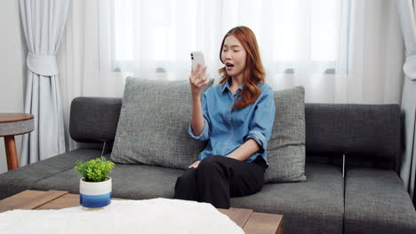 Charming-Asian-girl-sitting-on-the-couch-uses-video-calls-online-technology-with-a-friend-in-the-cozy-living-room-on-a-vacation-day