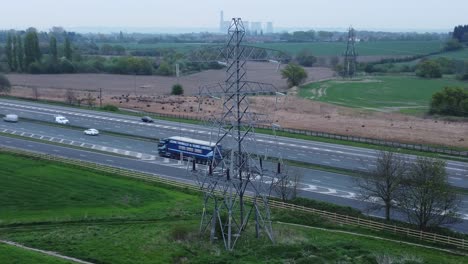 Vehicles-on-M62-motorway-passing-pylon-tower-on-countryside-farmland-fields-aerial-view-slow-push-in-left