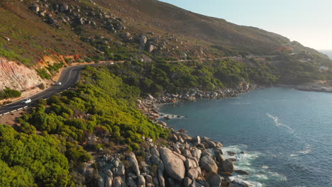 Cars-Driving-Through-Victoria-Road-In-Scenic-Seaside-Of-Oudekraal-Beach-In-South-Africa---aerial-shot