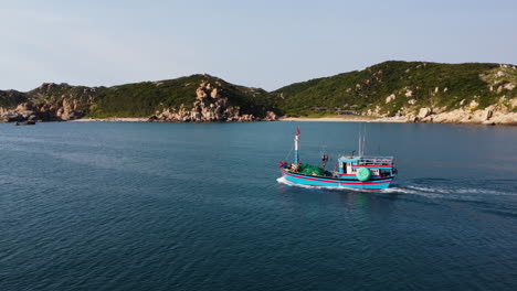 Colorful-fishing-vessel-leaving-tropical-port,-aerial-side-fly-view