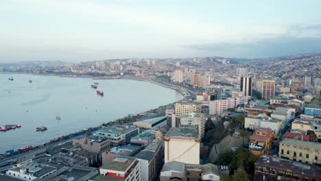 Aerial-dolly-in-of-Valparaiso-hillside-city-buildings-near-Sea-Port-and-ships-sailing-near-coastline-on-a-cloudy-day,-Chile