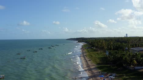 Dolly-in-aerial-drone-shot-of-the-beautiful-tropical-Penha-beach-coastline-near-the-capital-city-of-Joao-Pessoa-in-Paraiba,-Brazil-with-waves-crashing-into-the-sand-and-small-fishing-boats-docked