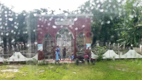 Four-young-Muslim-boys-huddle-together-for-shelter-from-a-sudden-downpour,-crouching-under-an-entrance-to-a-lush-green-park-as-raindrops-trickle-down-the-nearby-windowpanes