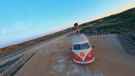 FPV-aerial-flying-around-a-woman-dancing-on-top-of-a-VW-minibus-parked-at-a-scenic-beach-at-sunset