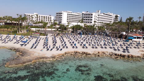 Aerial-towards-Nissi-Beach-resort-Sun-loungers-next-to-the-seashore,-Ayia-Napa-Cyprus