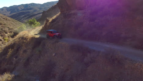 Red-Jeep-driving-on-narrow-hill-dirt-road-rising-dust-at-golden-hour,-aerial