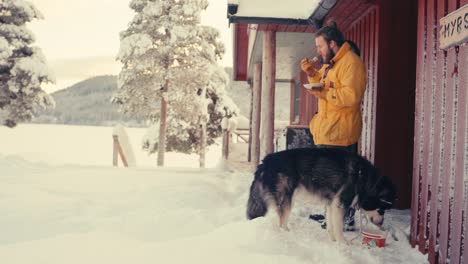 Man-And-Pet-Dog-Alaskan-Malamute-Eating-Meal-Outside-The-Cabin-On-A-Winter-Day