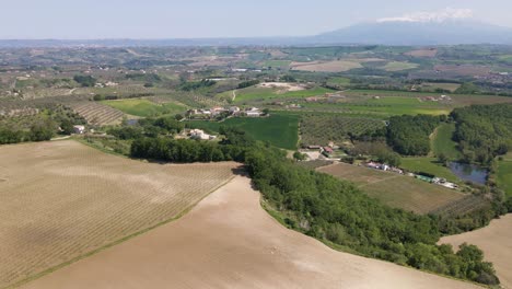 Wide-angle-drone-shot-panning-upwards-of-a-beautiful-vista-of-a-valley-with-farmland,-wineries,-medieval-villages-and-a-mountain-range-in-the-horizon-located-in-the-rural-Abruzzo-Italy-region